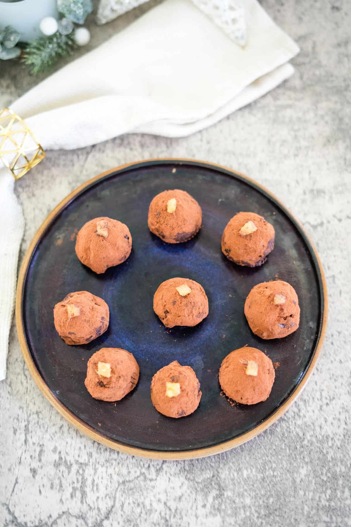 A black plate with ten round chocolate truffles, each topped with a small piece of candied ginger.