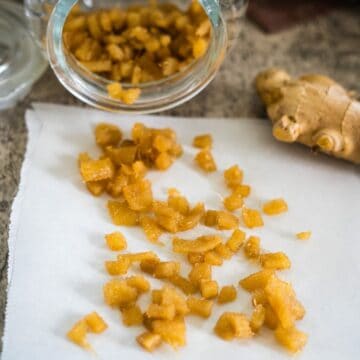 Chopped candied ginger pieces spill from a glass jar onto white parchment paper, with a fresh ginger root nearby on a countertop.