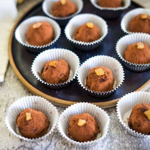 Chocolate truffles topped with small pieces of candied fruit are arranged in white paper cups on a round black tray.