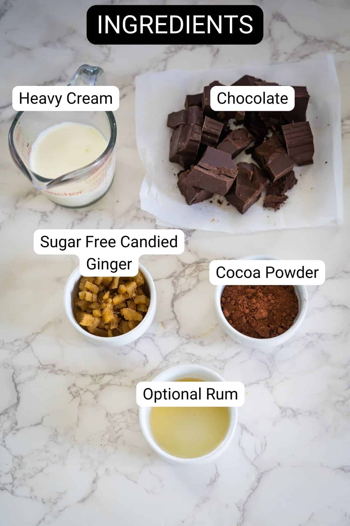 Ingredients for a recipe on a marble countertop include heavy cream, chocolate, sugar-free candied ginger, cocoa powder, and optional rum.