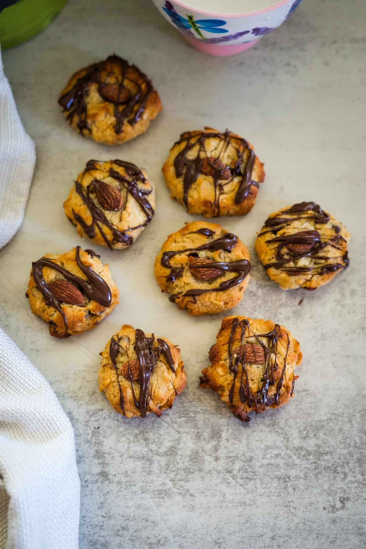 A group of keto almond joy cookies drizzled with chocolate are arranged on a gray surface. A white cloth and a floral cup are partially visible nearby.