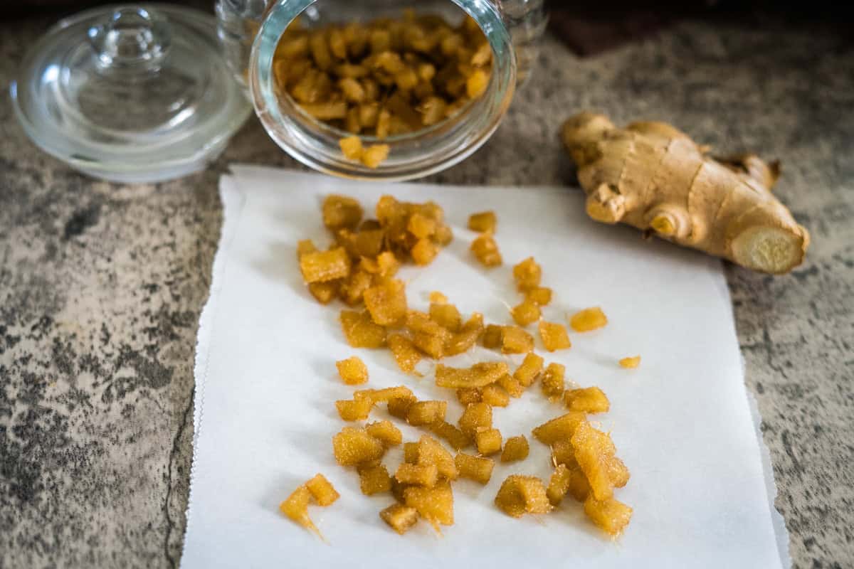 Sugar-free candied ginger pieces are scattered on white paper, next to a jar and a fresh ginger root on a textured surface.