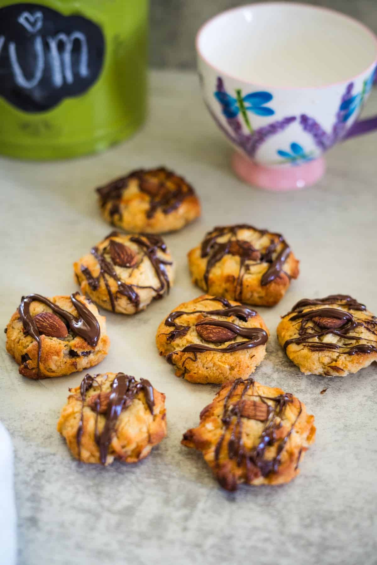 Chocolate-drizzled cookies with almonds are arranged on a table next to a decorative cup and a green container labeled "YUM.