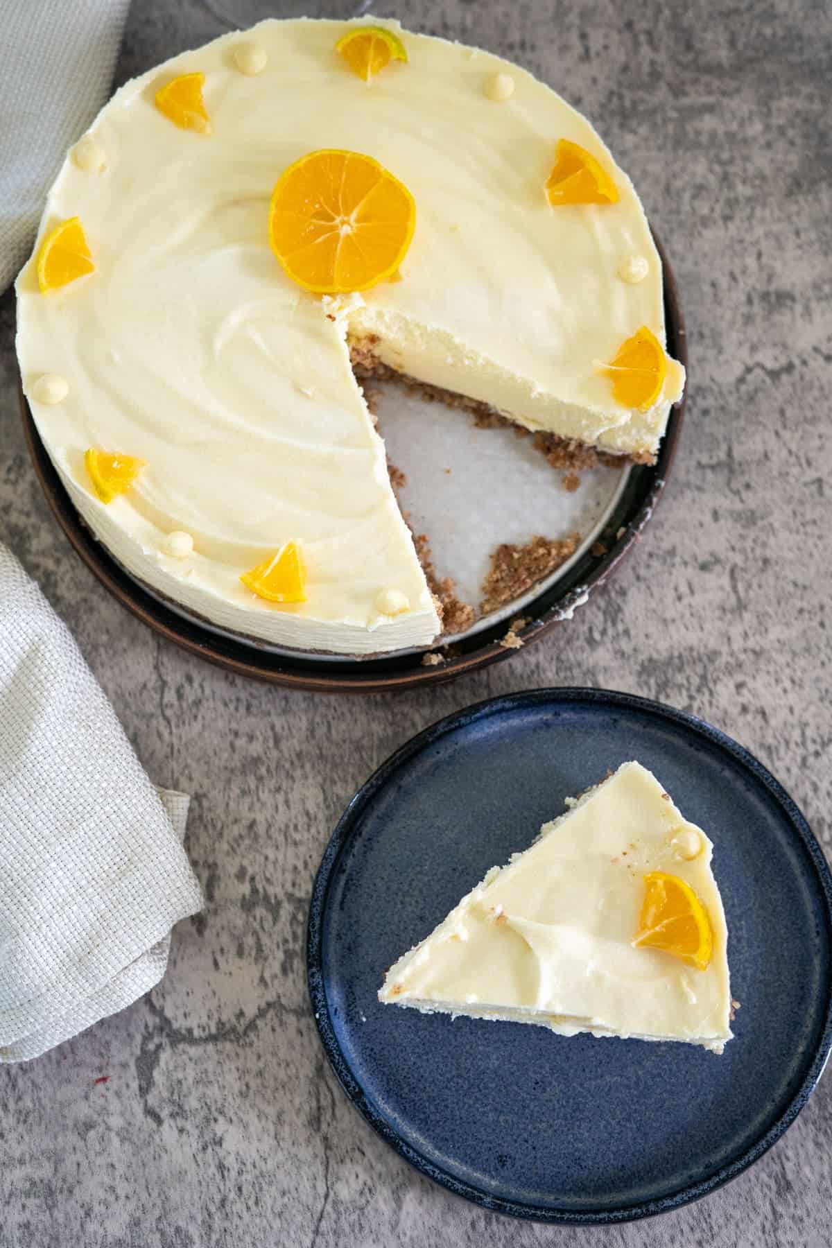 A cheesecake with a slice missing, topped with a whole orange slice in the center and small orange segments. A single slice is placed on a blue plate beside the cake.