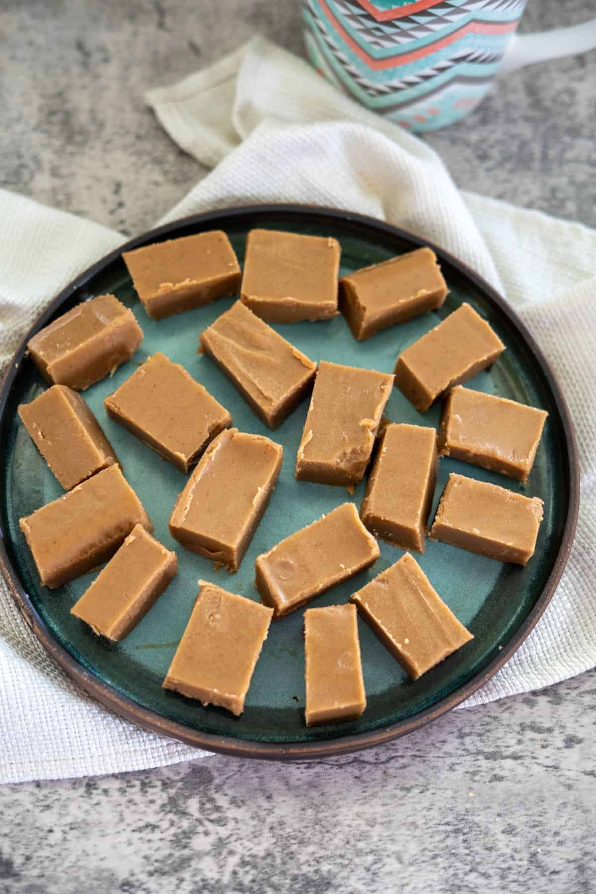 A round plate with rectangular pieces of light brown fudge arranged on a textured gray surface with a white cloth.