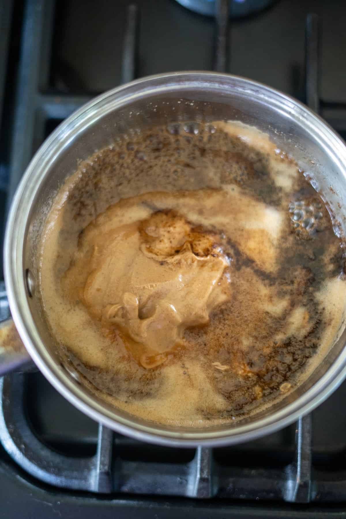 A saucepan on a stove contains bubbling brown liquid with a frothy texture, possibly coffee or sauce, evoking the rich aroma of keto peanut butter fudge.