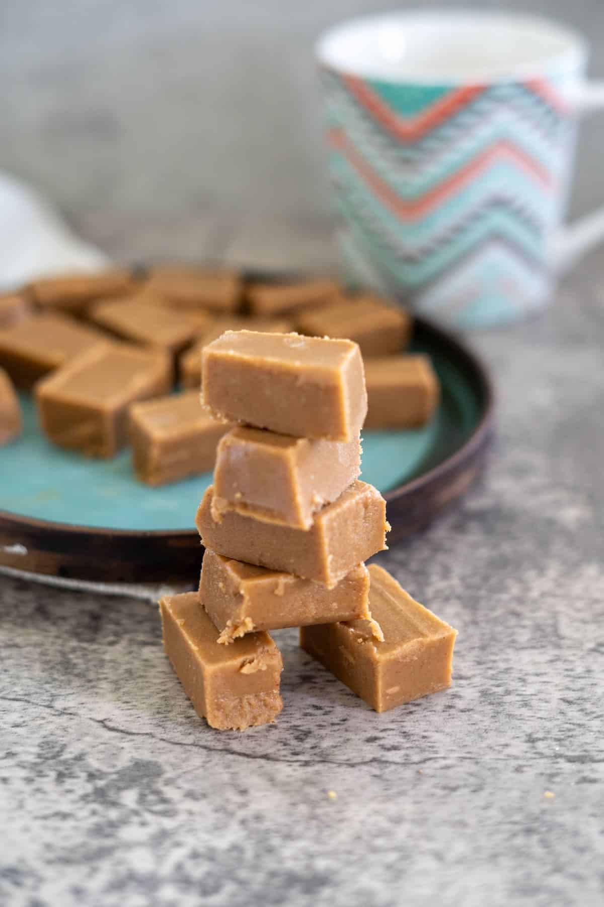 A stack of keto peanut butter fudge pieces is in the foreground, with more pieces on a blue plate in the background. A mug with a colorful zigzag pattern is partially visible.