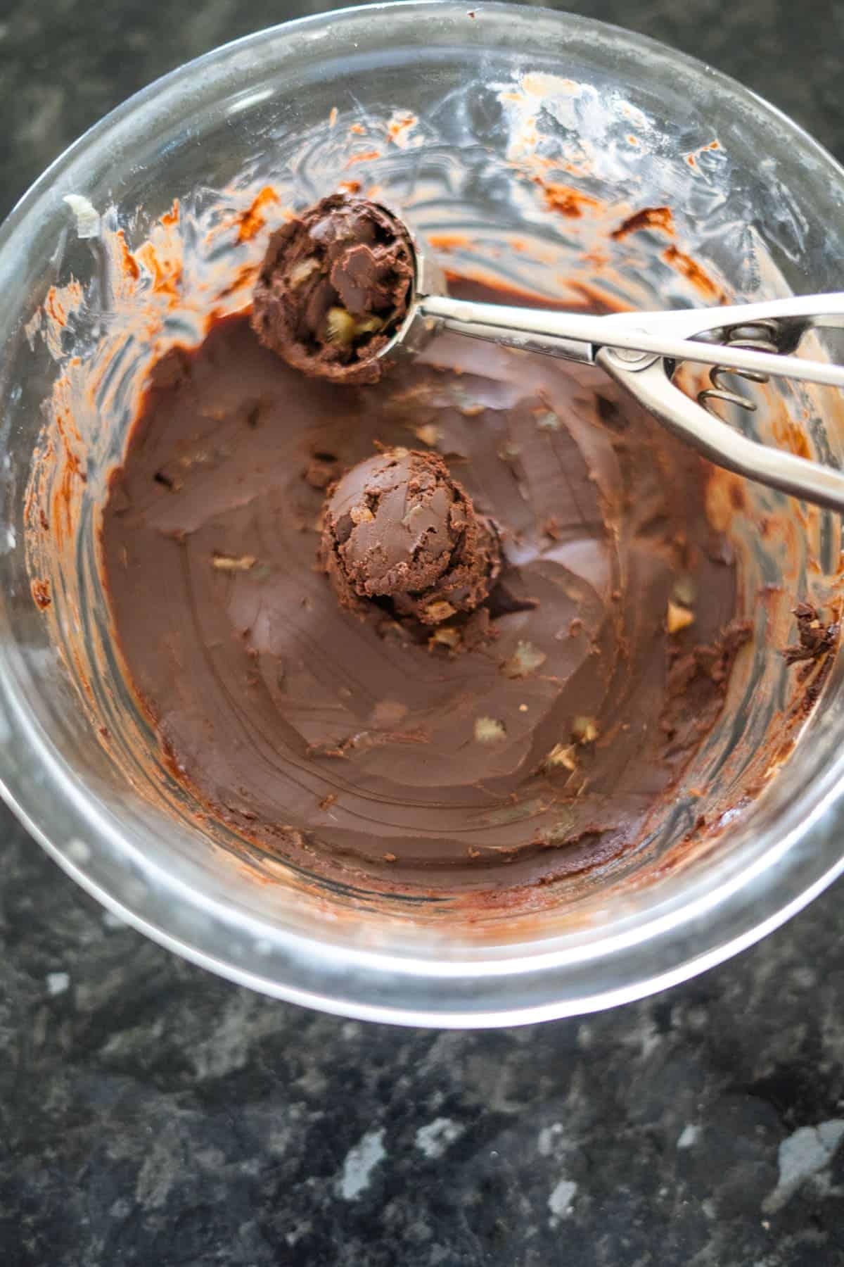 A glass bowl with chocolate cookie dough, a scoop of dough is being formed by a metal scooper.