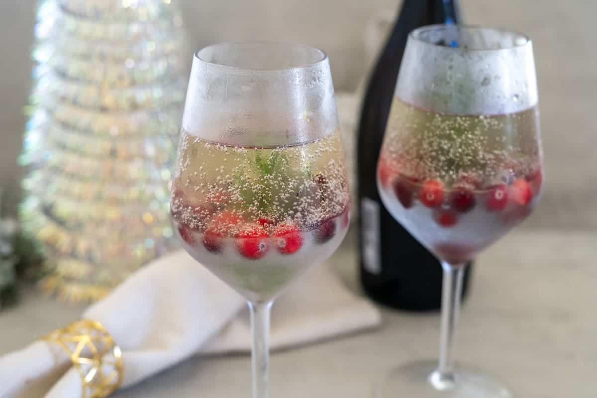 Two glasses of sparkling wine with cranberries and rosemary, reminiscent of a snow globe cocktail, sit next to a white napkin and a bottle in the background, framed by a festive backdrop.