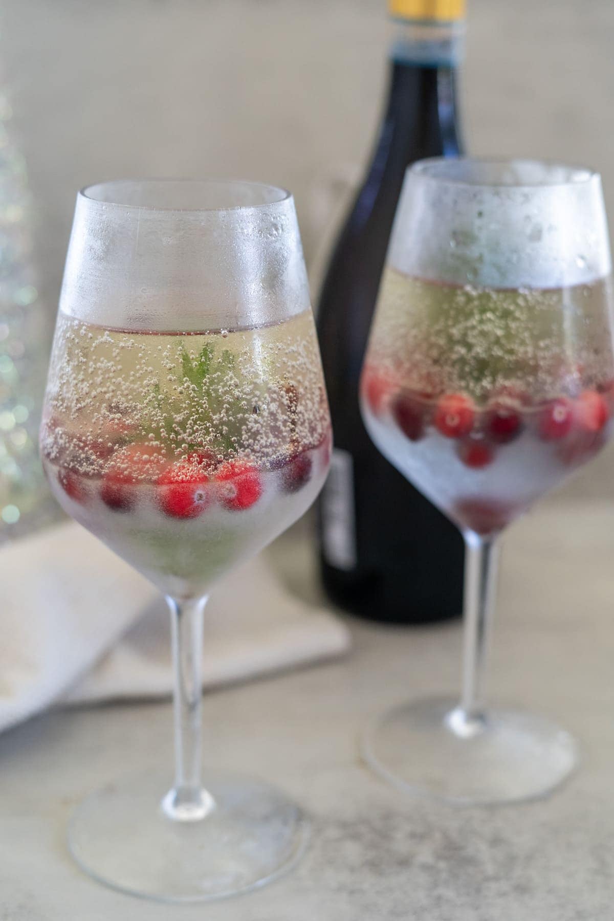 Two wine glasses filled with a sparkling snow globe cocktail, garnished with cranberries and a sprig of rosemary. A bottle is blurred in the background.