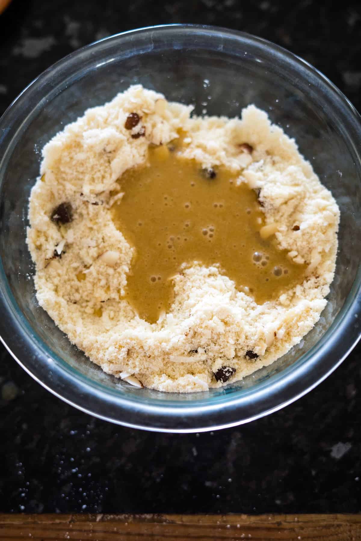 A glass bowl with flour mixture and raisins, with a pool of liquid in the center, on a dark countertop.