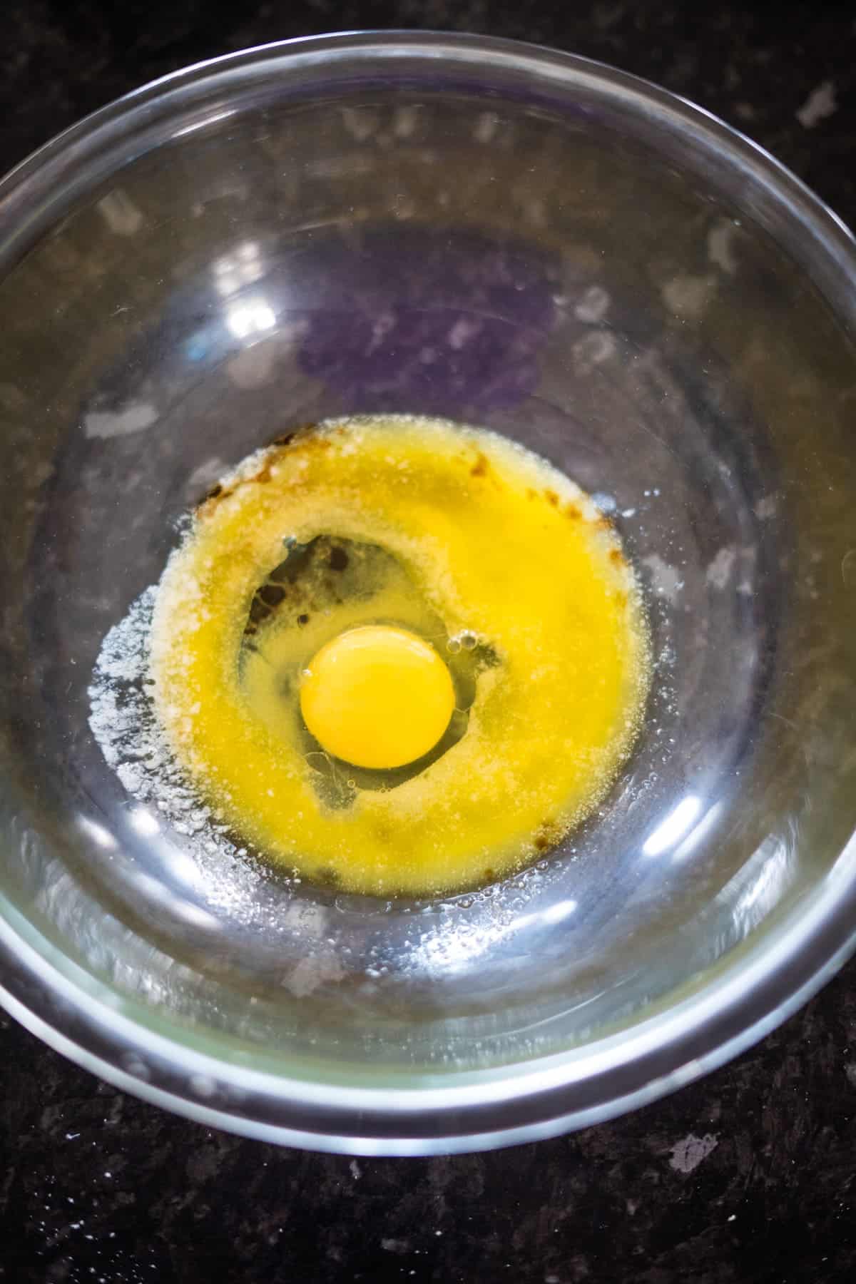 A cracked egg sits in a glass mixing bowl on a dark countertop.