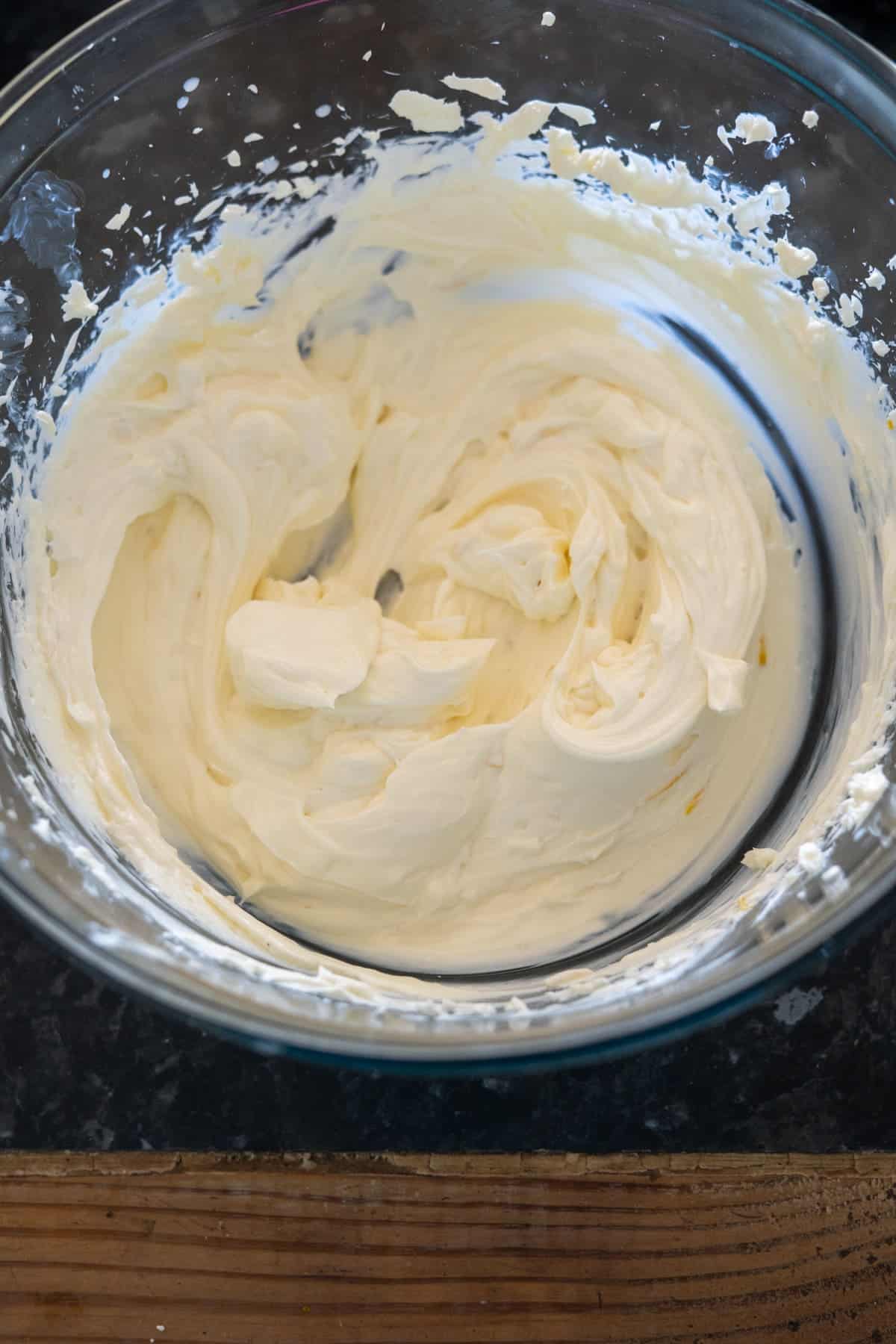 A glass bowl contains smooth, light yellow whipped cream on a dark countertop.