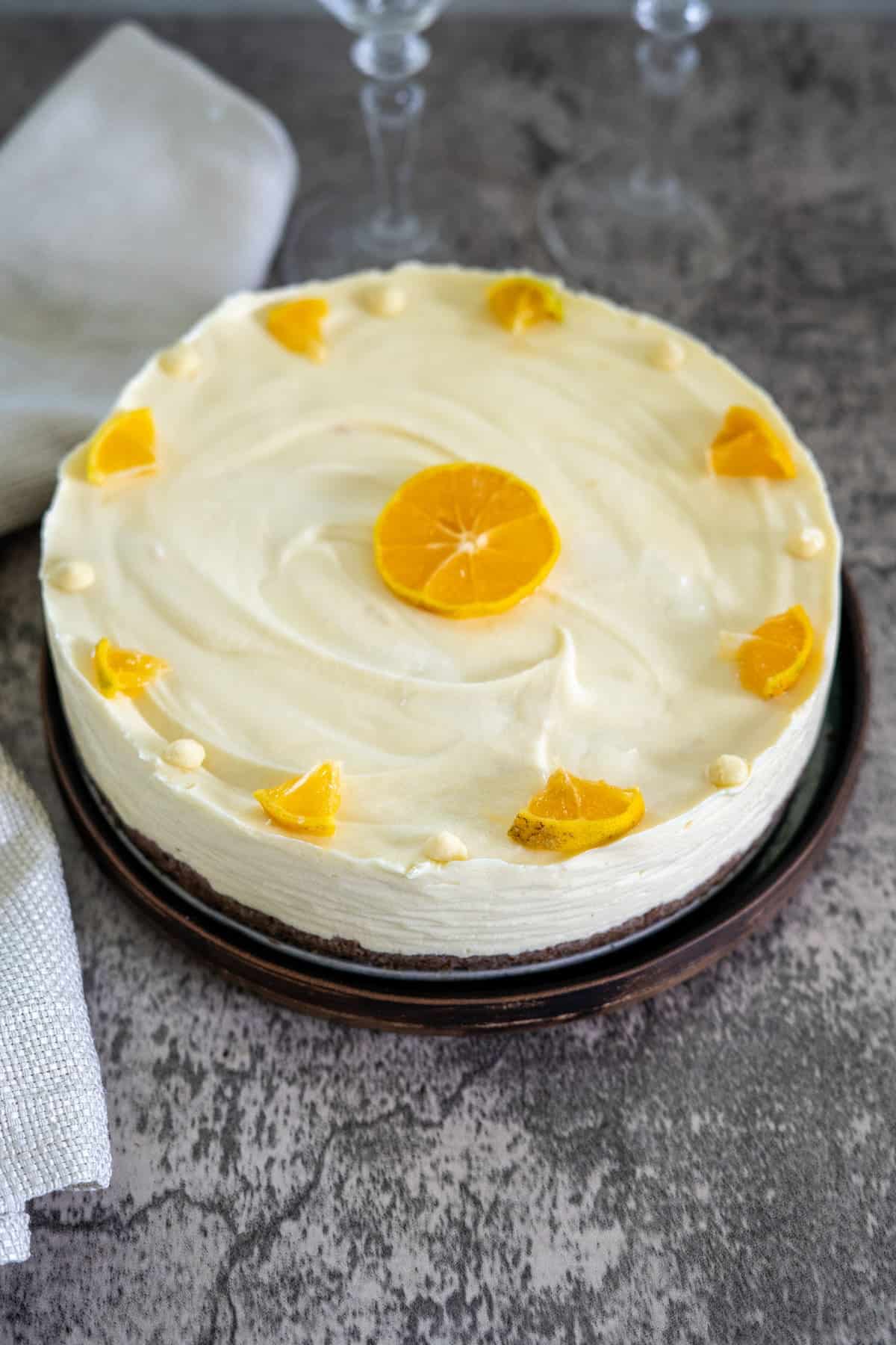 A round cheesecake topped with a slice of orange in the center and small orange segments around the edges, placed on a dark plate with a cloth napkin nearby.