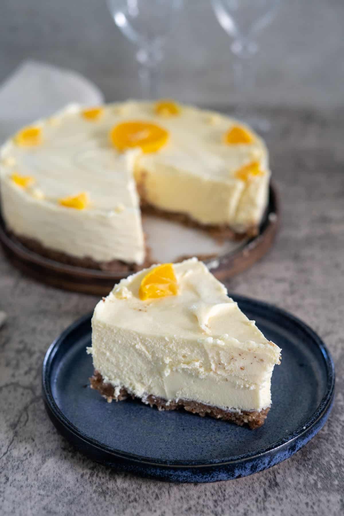 A slice of cheesecake with a crumbly base and creamy topping, garnished with peach pieces, is placed on a blue plate. The rest of the cheesecake is in the background.