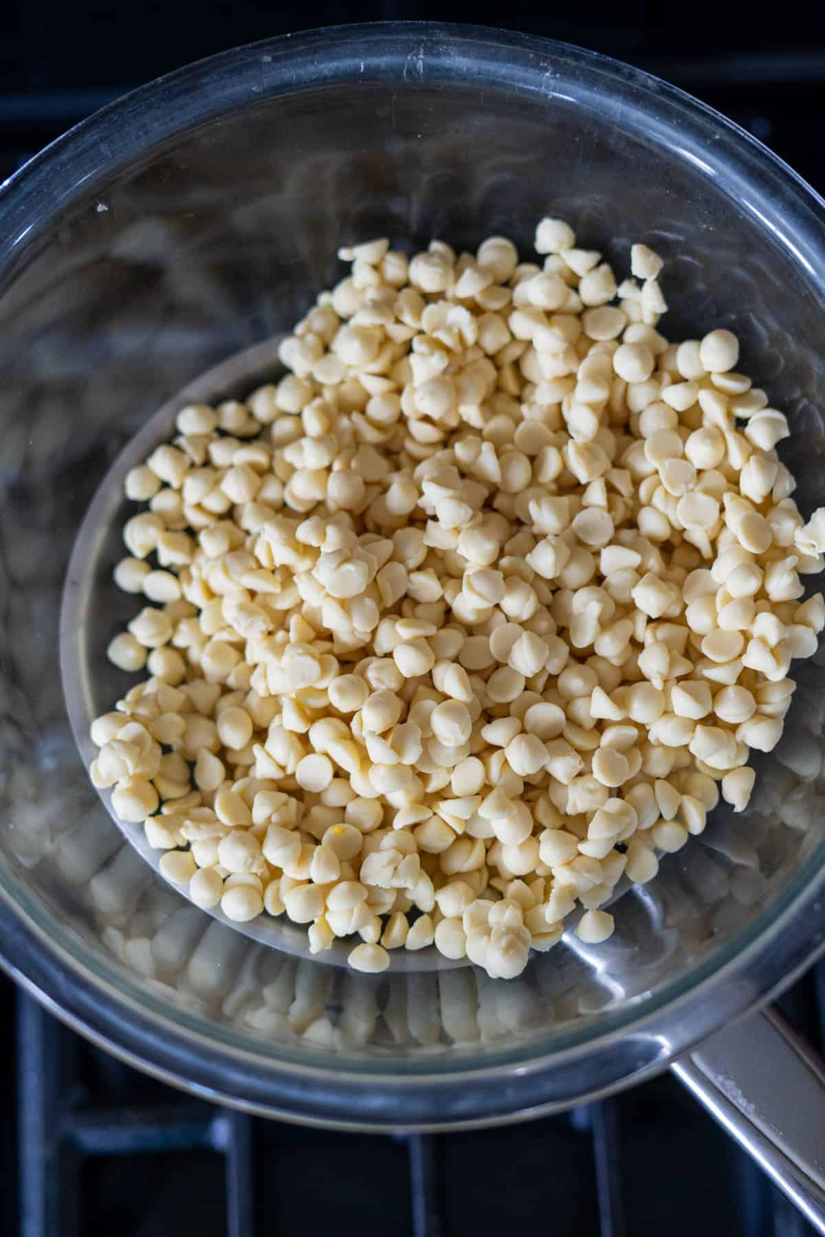 A glass bowl filled with white chocolate chips is placed over a pot on a stove, set up for a double boiler method.