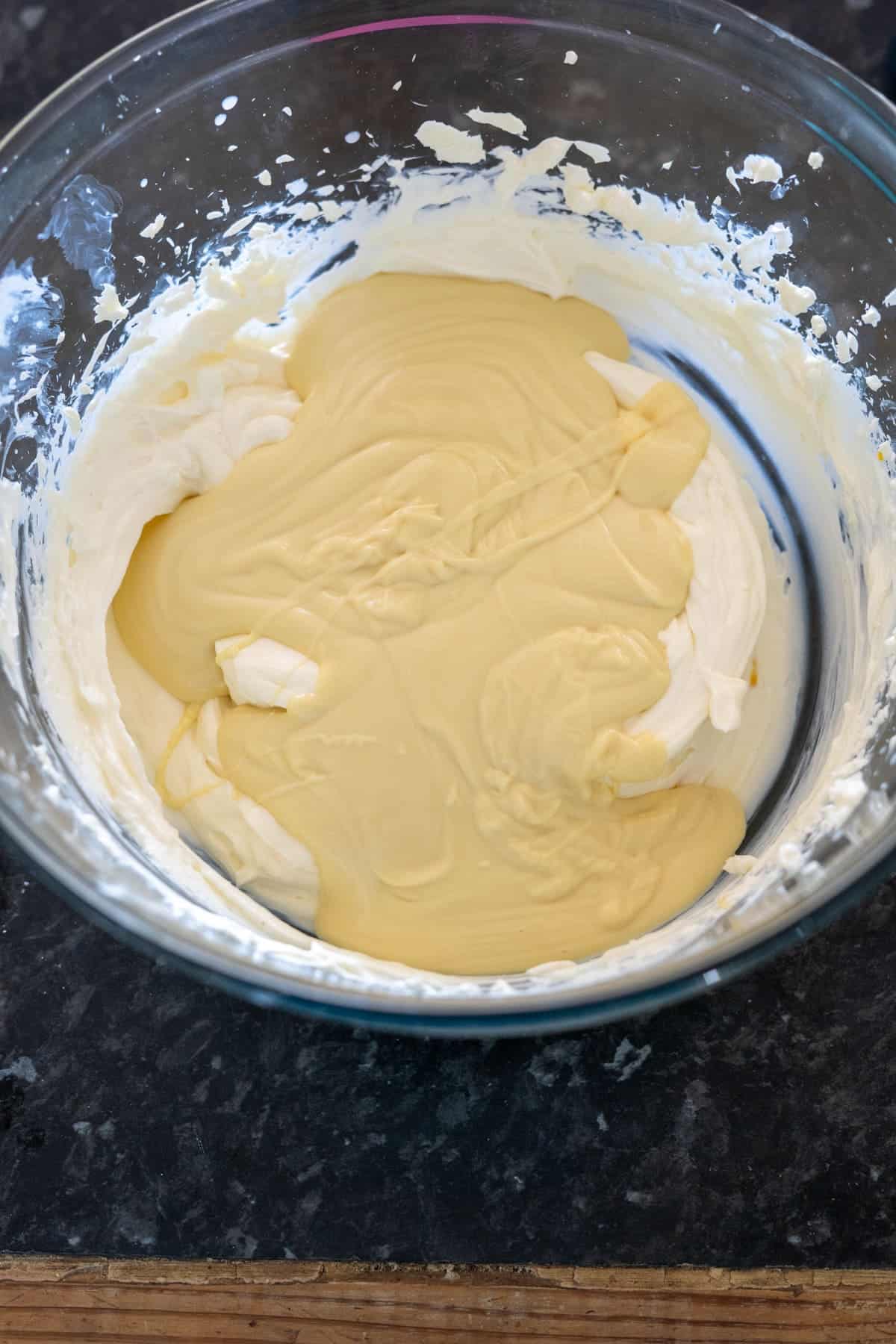 Glass bowl containing a mixture of whipped cream and yellow pudding on a dark countertop.